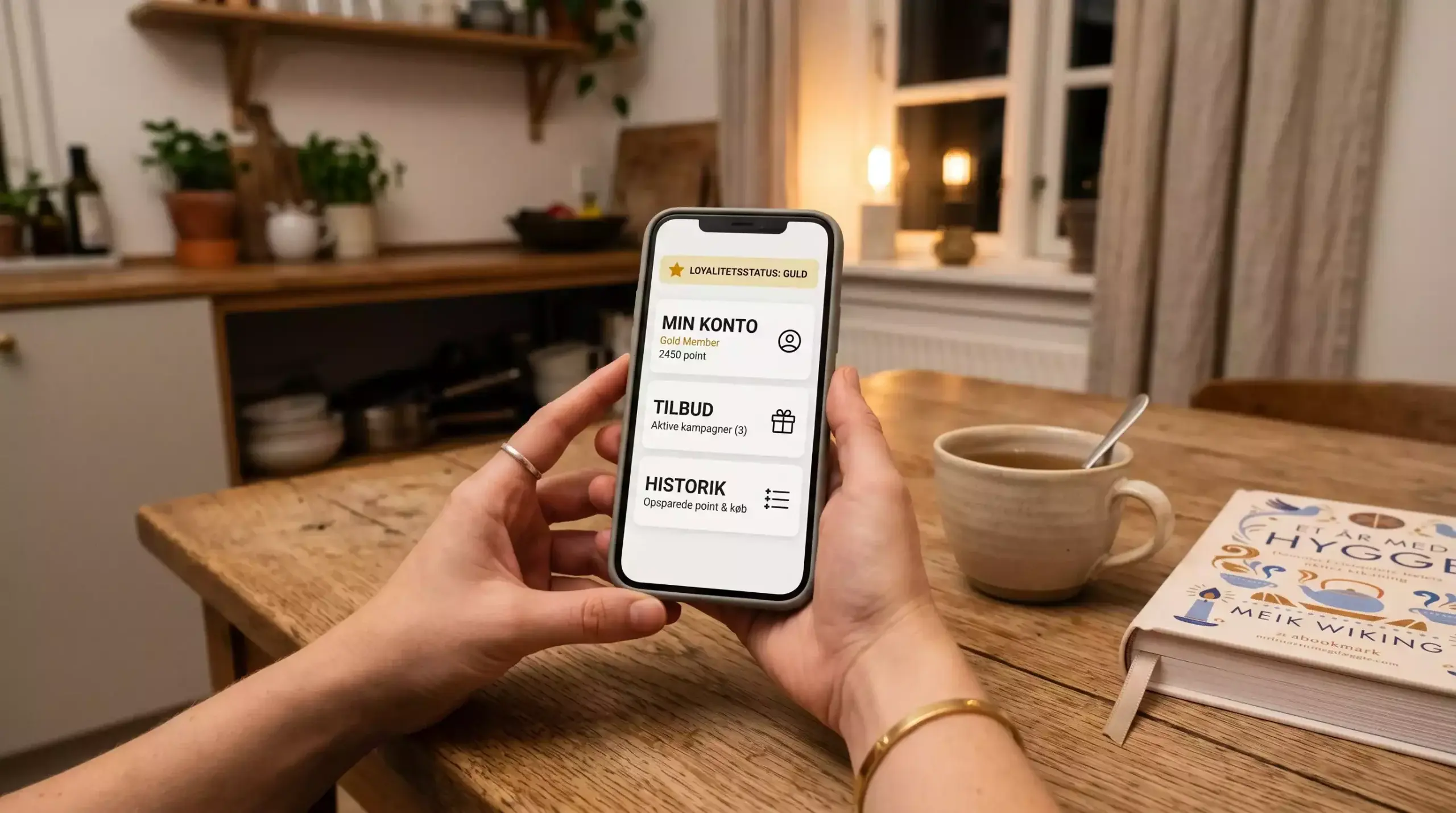 Smartphone med generisk kontooversigt på dansk holdt ved et hyggeligt køkkenbord