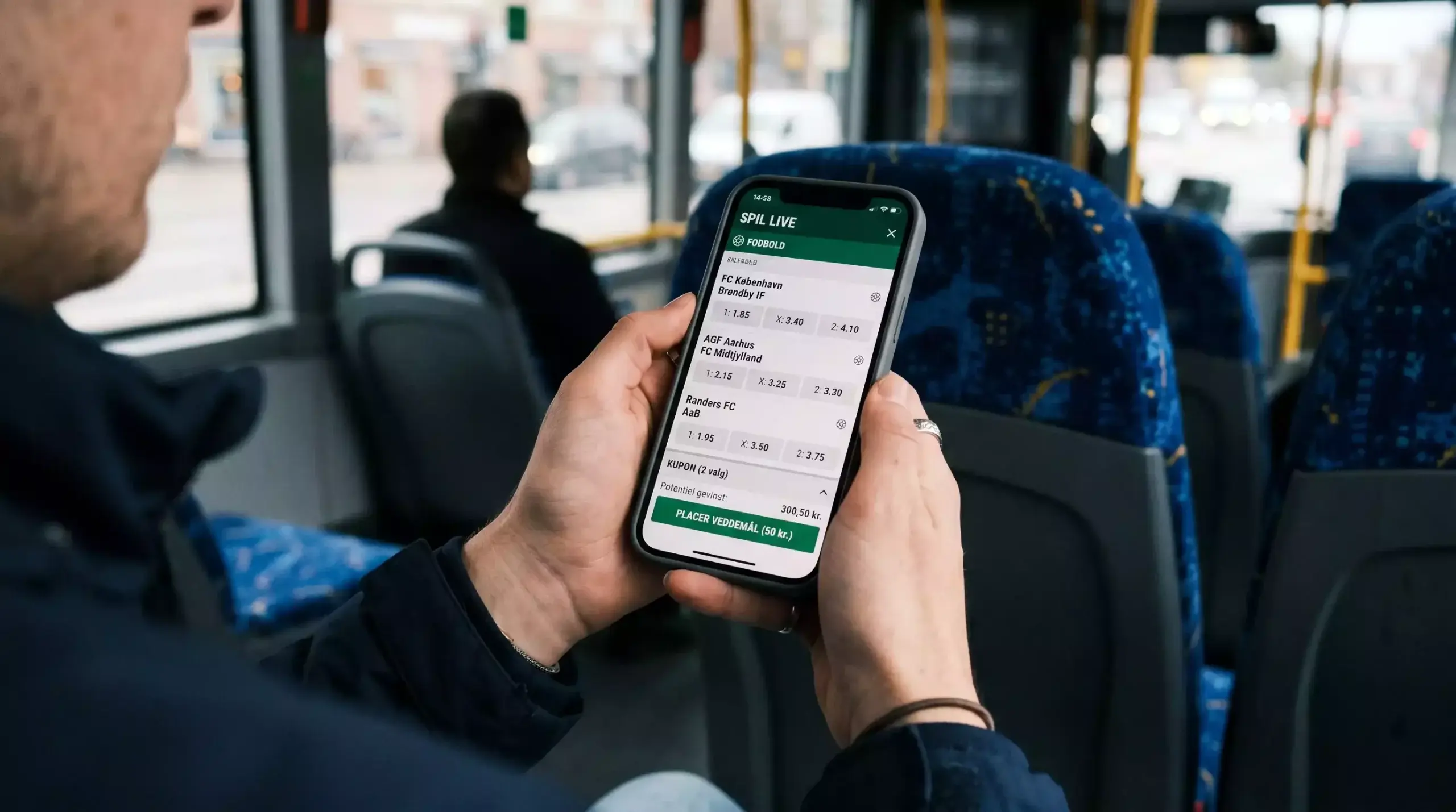 Smartphone med generisk sportsvæddemåls-app i dansk sprog holdt i hånden i en bus